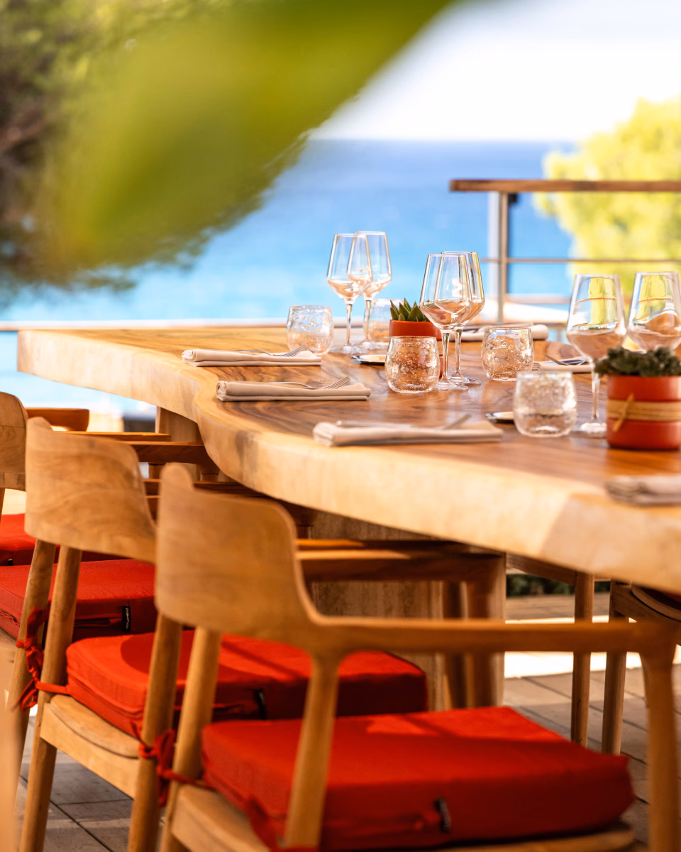 Table en bois avec des verres, des couverts et des serviettes, entourée de chaises en bois avec des coussins rouges, en extérieur près d'une étendue d'eau