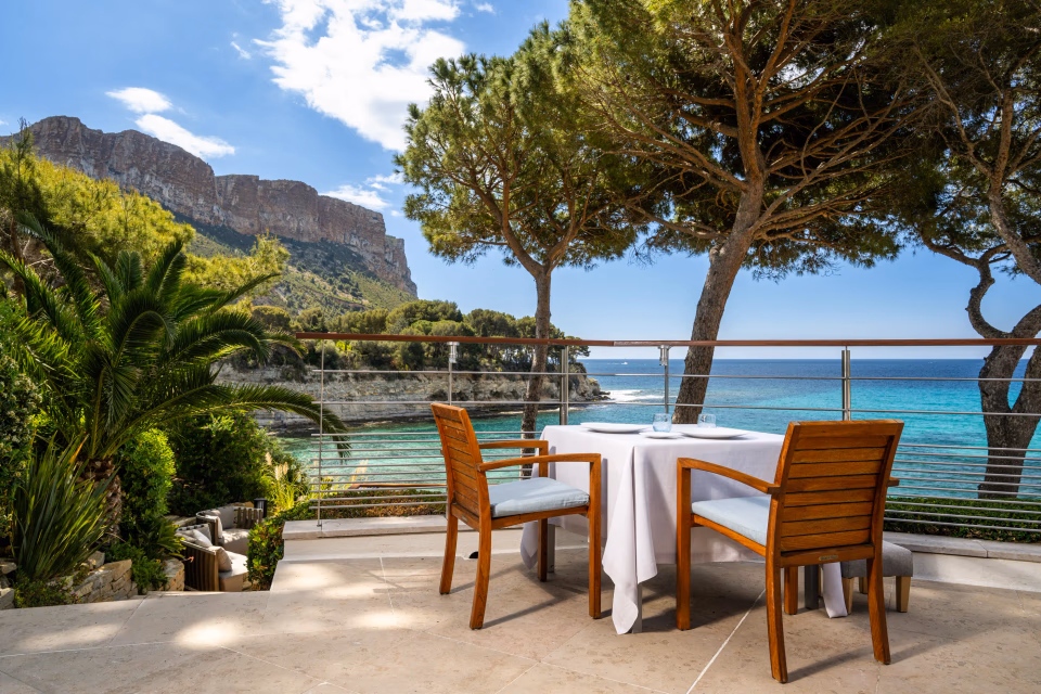 Table dressée avec deux chaises en bois sur une terrasse face à la mer, entourée d'arbres et de montagnes