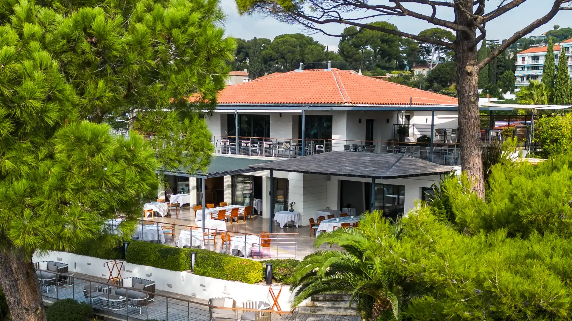 Restaurant avec terrasse et tables en extérieur, entouré d'arbres et de végétation, sous un ciel partiellement nuageux