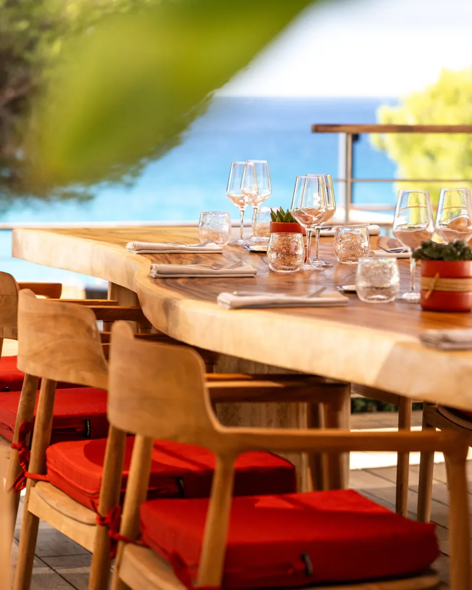 Table en bois avec des verres, des couverts et des serviettes, entourée de chaises en bois avec des coussins rouges, en extérieur près d'une étendue d'eau